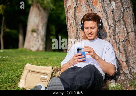 Un homme latino est assis sur l'herbe dans un parc, appuyé contre un arbre. Il se détend et se détend, en utilisant son smartphone pour se déconnecter du stress quotidien. Banque D'Images