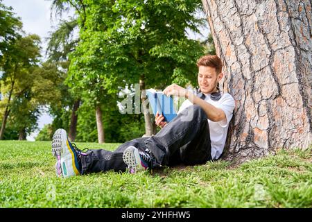 Un homme est assis sur l'herbe dans un parc, appuyé contre un arbre tout en étant absorbé par la lecture d'un livre, profitant d'un moment de tranquillité dans la nature. Banque D'Images