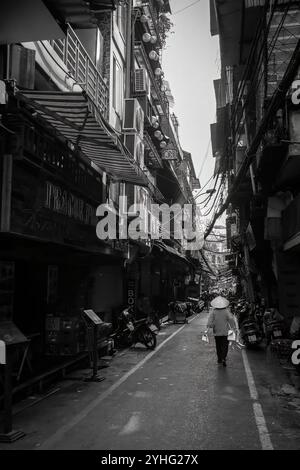 Une femme avec un chapeau conique vietnamien traditionnel marche dans une rue étroite de Hanoi, en passant devant des bâtiments et des motos très bien remplis. Banque D'Images