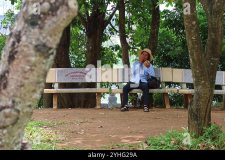 Un homme se repose sur un banc à l'ombre des arbres, dégustant une collation dans un cadre paisible, portant un chapeau à larges bords et un badge d'identification. Banque D'Images