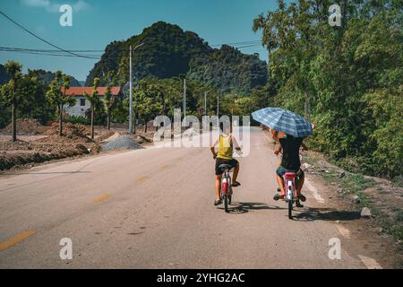 Deux garçons montent à vélo sur une route rurale au Vietnam, l'un tenant un grand parapluie pour l'ombre. La scène capture une journée ensoleillée au milieu d'une vue panoramique sur la montagne. Banque D'Images