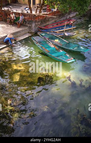 Une scène paisible au bord de la rivière au Vietnam avec des bateaux traditionnels amarrés, un homme se lavant au bord de l'eau, et les habitants se relaxant à proximité, mettant en valeur la vie quotidienne. Banque D'Images