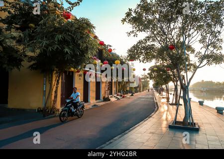 Un homme du coin roule à moto le long d'une rue ornée de lanternes au bord de la rivière à Hoi an, au Vietnam, capturant l'ambiance sereine du petit matin. Banque D'Images