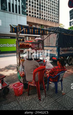 Deux femmes assises à un stand de nourriture de rue au Vietnam, partageant un moment de calme au milieu de la vie de rue animée de la ville. Banque D'Images