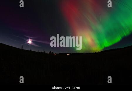 Les aurores boréales dansent à travers le ciel au-dessus d'une ferme rurale, avec des lignes électriques qui s'étendent à travers le paysage, la pleine lune brille, et un satellite Banque D'Images
