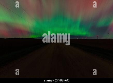 L'aurora borealis brille au-dessus d'une route de campagne tranquille, avec un arbre solitaire debout au bord de la route, des moulins à vent au loin, et un passage satellite t Banque D'Images