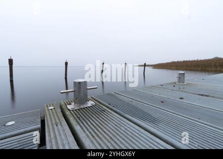 Une vue sereine de novembre sur la lagune de Szczecin, mettant en valeur une jetée en bois tranquille s'étendant dans les eaux calmes et brumeuses avec des poteaux d'amarrage. Banque D'Images