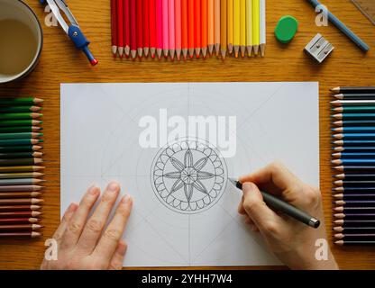 Mains d'une femme caucasienne dessinant un motif mandala sur un papier blanc couché sur une table en bois, dessinant des ustensiles et une tasse avec du café à proximité Banque D'Images