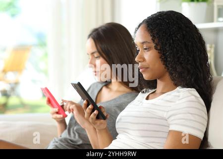 Profil de deux amis sérieux ou colocataires s'ignorant l'un l'autre en utilisant leur téléphone à la maison Banque D'Images