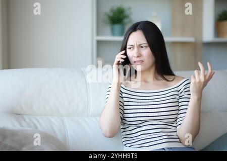Femme asiatique en colère parlant au téléphone assis sur un canapé à la maison Banque D'Images