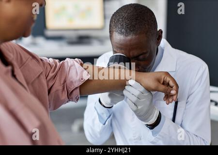 Vue de face portrait d'un médecin afro-américain regardant dans dermascope tout en examinant le patient à la clinique de dermatologie espace de copie Banque D'Images