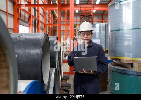 Un homme portant un casque et une combinaison mécanique de protection contre la poussière vérifie l'ordinateur portable dans l'usine de tôle. La pile de rouleaux en tôle d'acier est dans le Banque D'Images