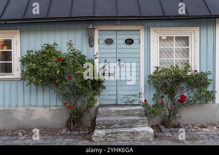 Une maison en bois bleu clair avec une vieille porte flanquée de roses rouges dans un cadre confortable et rustique, ville hanséatique de Visby, site du patrimoine mondial de l'UNESCO, Gotlan Banque D'Images