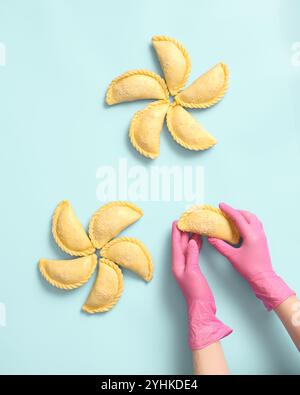 Vue de dessus des mains d'une boulangère dans des gants en caoutchouc rose déposant des tartes crues sur un fond bleu, produits semi-finis congelés Banque D'Images