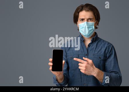 Homme dans masque de protection pointant vers le smartphone avec écran noir Banque D'Images