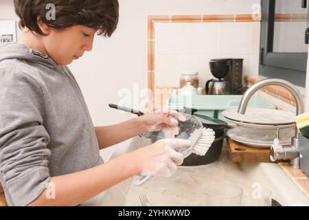 Un enfant lave la vaisselle avec une brosse dans une cuisine. Le garçon porte un sweat à capuche gris et utilise une brosse à vaisselle pour nettoyer un verre. La cuisine est encombrée Banque D'Images