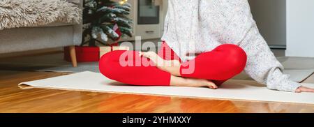 Yoga et méditation pendant les vacances de Noël à la maison. Gros plan des jambes d’une femme en collant de sport rouge en position demi-lotus. Banque D'Images