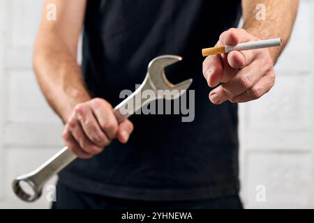 Bavière, Allemagne - 12 novembre 2024 : artisan avec clé, outil et cigarette à la main. Image symbolique pour fumer au travail, ou la consommation de cigarettes pendant le travail, les heures de travail. *** Handwerker mit Schraubenschlüssel, Werkzeug und Zigarette in der hand. Symbolbild für Rauchen am Arbeitsplatz, bzw. Konsum von Zigaretten während der Arbeit, Arbeitszeit. Banque D'Images