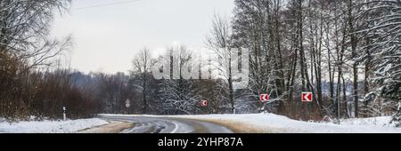 Une vue panoramique sur une route d'hiver en virage brusque, entourée de paysages glacés. Des panneaux de signalisation routière sont visibles sur le côté, avertissant de conditions dangereuses Banque D'Images