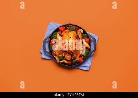 Vue de dessus avec poulet rôti et légumes dans une fonte de fer, fraîchement cuit, minimaliste sur une table orange Banque D'Images