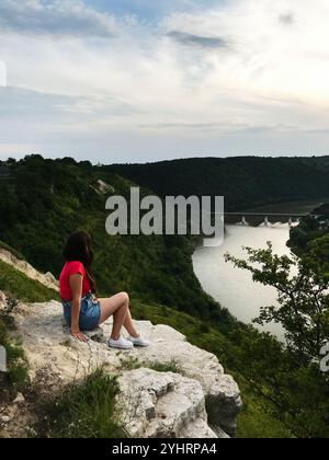 Une femme en tenue décontractée est assise sur le bord d'une falaise, regardant au-dessus d'une belle vallée de rivière. Le paysage est luxuriant de verdure et le ciel est couvert, créa Banque D'Images