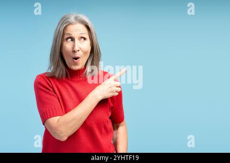 Portrait de femme mature excitée et heureuse portant un pull rouge pointant le doigt sur l'espace de copie, isolé sur fond bleu. Concept de publicité Banque D'Images