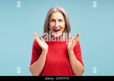 Portrait d'une femme âgée excitée et ravie avec la bouche ouverte portant un pull rouge, regardant la caméra, posant isolé sur fond bleu. Publicité Banque D'Images
