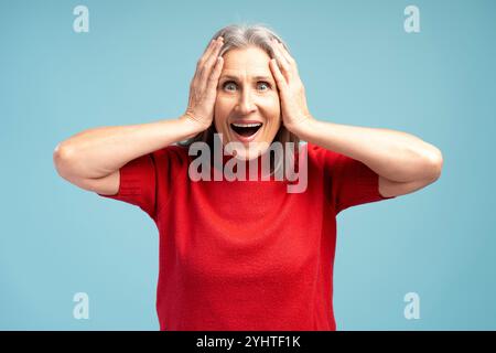 Femme mature excitée et positive portant un pull rouge avec la bouche ouverte regardant la caméra, se tenant isolée sur fond bleu. Shopping, concept de vente Banque D'Images