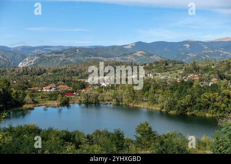 Vue panoramique sur un lac serein près de Petrosani, Roumanie, entouré de verdure luxuriante et de montagnes lointaines sous un ciel bleu clair. Banque D'Images