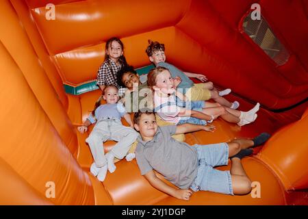 Grand groupe de mignons enfants interculturels amicaux regardant la caméra tout en étant couchés les uns à côté des autres par glissière en cuir Banque D'Images