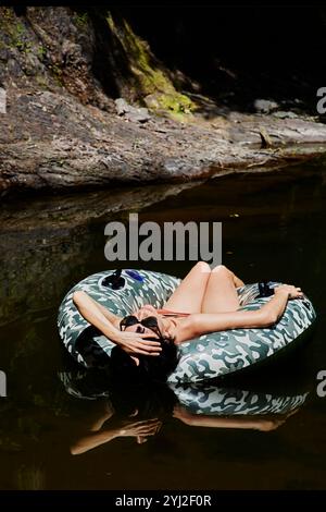 Femme se relaxant sur un anneau gonflable dans une rivière de forêt sereine, se prélassant dans la lumière du soleil avec ses mains derrière sa tête. Banque D'Images