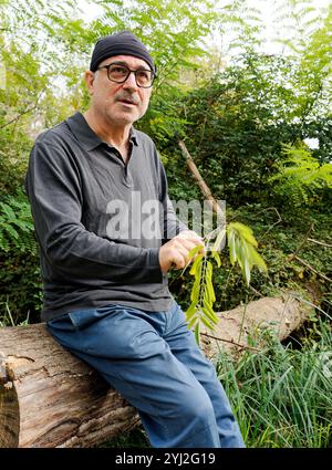 Homme mûr avec des lunettes et un bonnet assis sur une bûche dans un jardin verdoyant préparant une branche de plante pour la pêche, Italie Banque D'Images