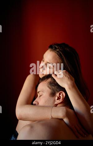 Une femme embrasse tendrement un homme par derrière, les deux avec des expressions introspectives sur un fond chaud et rouge. Banque D'Images