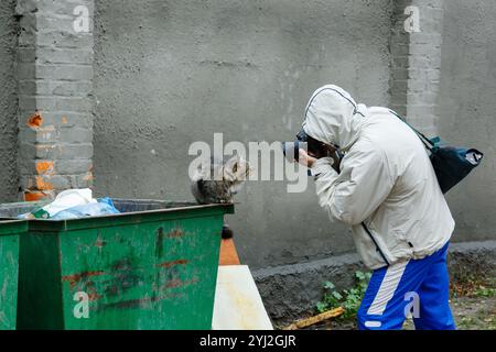 Un photographe prend une photo d'un chat. Photographe masculin tenant un appareil photo prenant des photos d'un chat errant assis sur une poubelle un jour de pluie. Banque D'Images