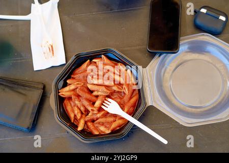 Pâtes Penne avec sauce tomate dans un récipient noir à emporter à côté d'une fourchette en plastique blanc, téléphone portable, étui d'écouteurs sans fil, portefeuille Banque D'Images