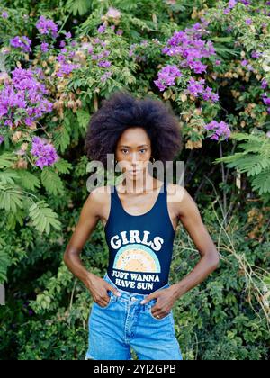 Femme confiante avec un afro saisissant se tient devant un buisson de fleurs violettes, portant un débardeur et un Jean bleu. Banque D'Images