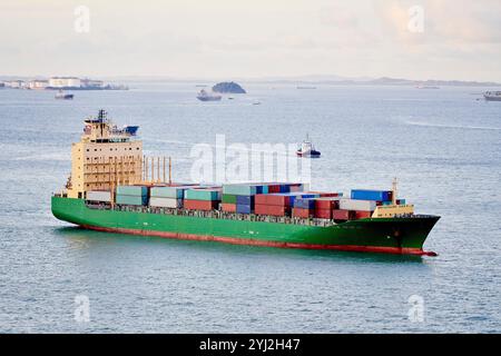 Cargo cargo chargé de conteneurs colorés guidé par un remorqueur sur une mer calme avec d'autres bateaux en arrière-plan, Singapour Banque D'Images