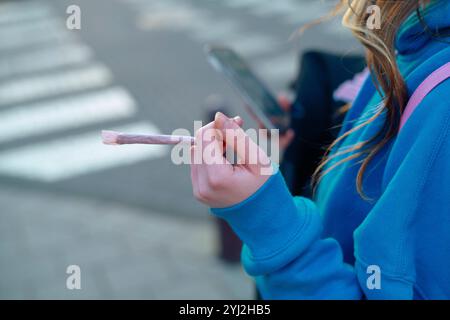 Gros plan de la main d'une personne tenant une cigarette ou un joint tout en utilisant un smartphone, portant un sweat à capuche bleu. Banque D'Images