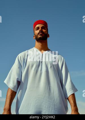 Homme confiant debout contre un ciel bleu clair portant une tunique blanche et un bandeau rouge, Maroc Banque D'Images