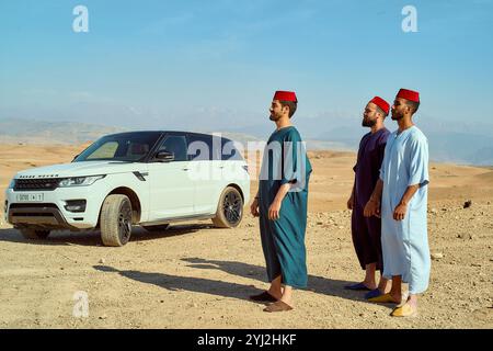 Quatre hommes en tenue traditionnelle se tiennent debout à côté d'un SUV blanc dans un paysage désertique, Maroc Banque D'Images