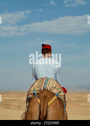 Un homme portant un bandeau rouge et une chemise blanche monte à dos de chameau dans le désert sous un ciel bleu clair, le Maroc Banque D'Images