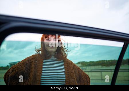 Une femme souriante aux cheveux roux vue à travers une fenêtre de voiture ouverte sur un fond de ciel nuageux. Banque D'Images