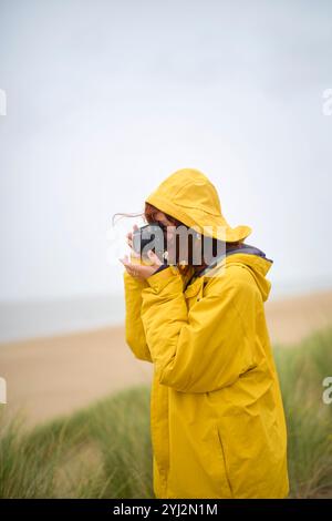 Un photographe dans un imperméable jaune prend une photo sur une plage au ciel couvert, Belgique Banque D'Images