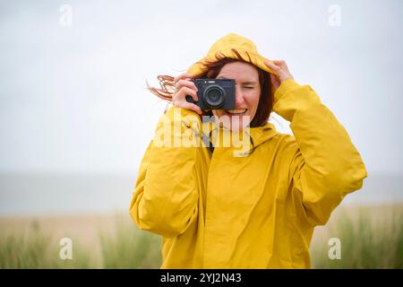 Femme dans un imperméable jaune prend une photo avec un appareil photo numérique un jour venteux au bord de la mer, Belgique Banque D'Images