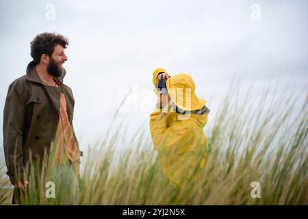 Un adulte dans un imperméable jaune prend une photo d'un homme barbu portant une veste verte, tous deux debout au milieu d'herbes hautes sous un ciel couvert, Belgique Banque D'Images