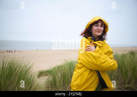 Une femme en veste jaune se tient près de l'herbe de plage sur une plage de sable sous un ciel couvert, Belgique Banque D'Images
