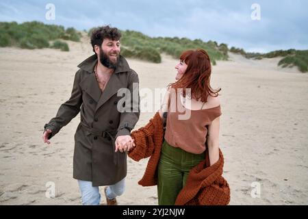 Couple joyeux se tenant la main et riant en marchant sur une plage de sable sous un ciel nuageux, Belgique Banque D'Images