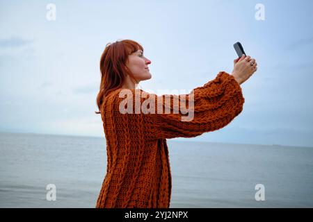 Femme dans un pull orange brûlé prend un selfie au bord de la mer sous un ciel couvert, Belgique Banque D'Images