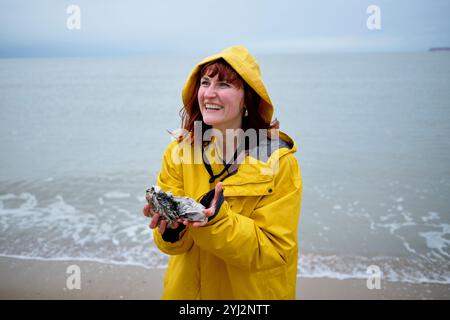 Femme souriante en imperméable jaune tenant une huître à la plage un jour couvert, Belgique Banque D'Images