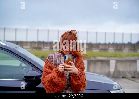 Femme dans un pull orange avec plaisir en utilisant son smartphone à côté d'une voiture avec un fond flou, Belgique Banque D'Images